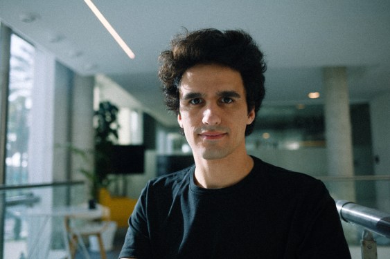 A man wearing a large black t-shirt stands in a sunlit office.